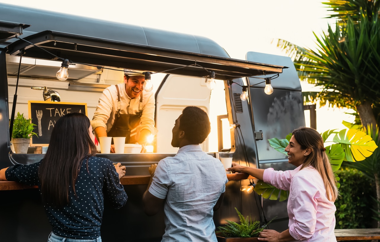 Three patrons smile & laugh at a food truck operator in a glossy, black mobile kitchen